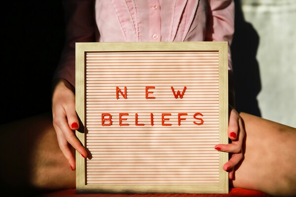 Jeune femme aux ongles vernis rouges tenant un cadre avec l'inscription "New Beliefs" en lettrage rouge