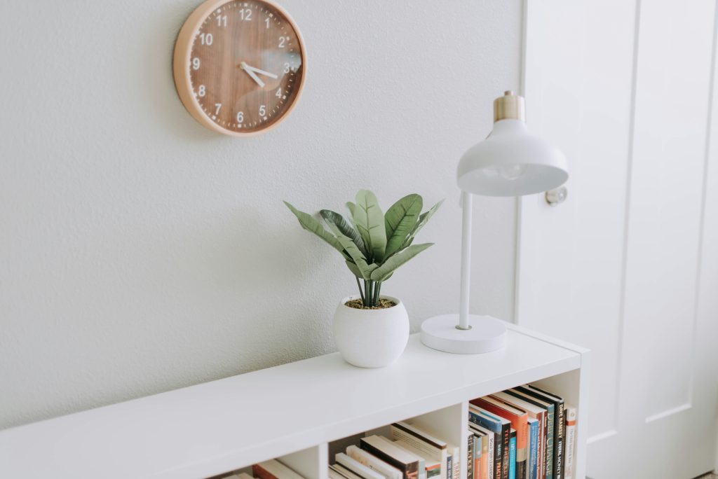 Intérieur de style minimaliste avec horloge murale en bois, petite plante verte en pot blanc, lampe blanche et étagère remplie de livres. Une inspiration au minimalisme.