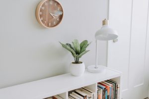 Intérieur de style minimaliste avec horloge murale en bois, petite plante verte en pot blanc, lampe blanche et étagère remplie de livres. Une inspiration au minimalisme.