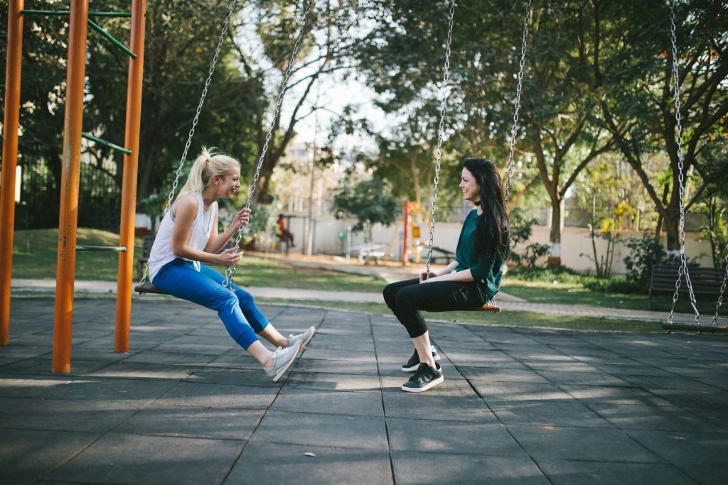 Deux amies souriantes en tenue de sport en pleine discussion sur des balançoires en léger mouvement dans un parc ensoleillé. Évocateur des bienfaits de bouger.