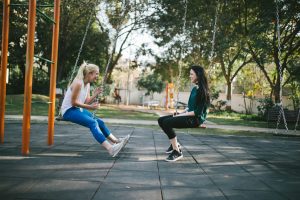 Deux amies souriantes en tenue de sport en pleine discussion sur des balançoires en léger mouvement dans un parc ensoleillé. Évocateur des bienfaits de bouger.