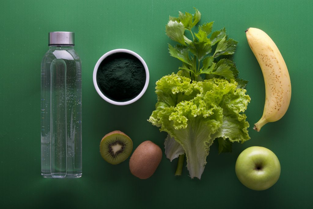 Bouteille d’eau, kiwi, pomme verte, banane, laitue et poudre verte sur fond vert – aliments frais pour une alimentation saine et détox