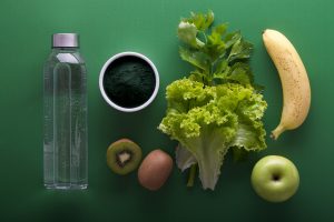 Bouteille d’eau, kiwi, pomme verte, banane, laitue et poudre verte sur fond vert – aliments frais pour une alimentation saine et détox