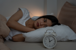 Une personne allongée dans son lit cherchant le sommeil, les yeux ouverts et l’air préoccupé, regardant un réveil analogique posé sur la table de chevet dans une chambre sombre. Photo pour illustrer l'article "Sommeil: et s'il n'était pas le problème?"