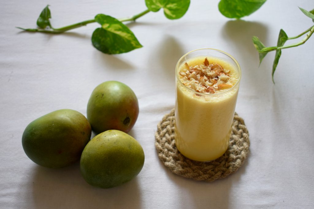 Verre de smoothie jaune à base de fruits, garni d’amandes, posé sur un sous-verre, avec des mangues fraîches à côté. Photo utilisée pour illustrer l'article Les faux-amis de l’alimentation saine
