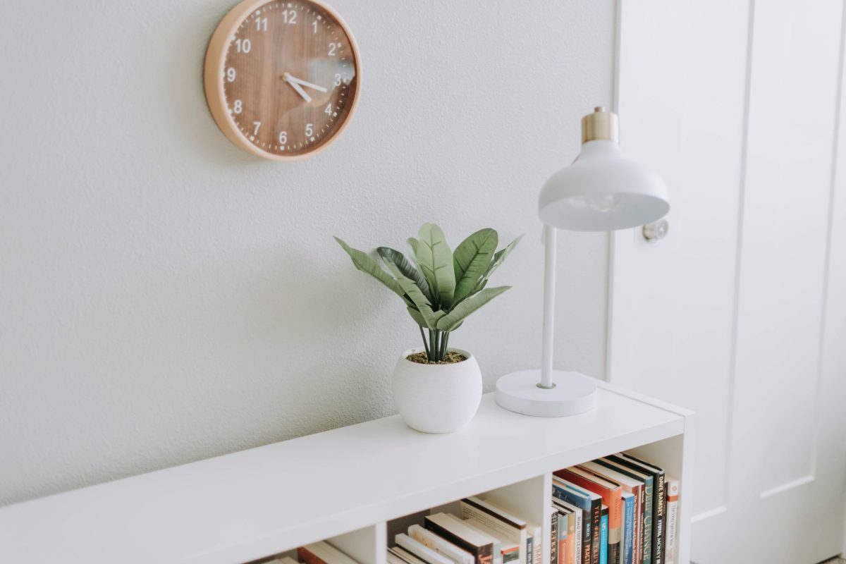 Intérieur de style minimaliste avec horloge murale en bois, petite plante verte en pot blanc, lampe blanche et étagère remplie de livres. Une inspiration au minimalisme.