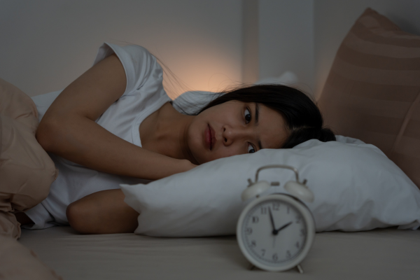 Une personne allongée dans son lit cherchant le sommeil, les yeux ouverts et l’air préoccupé, regardant un réveil analogique posé sur la table de chevet dans une chambre sombre. Photo pour illustrer l'article "Sommeil: et s'il n'était pas le problème?"