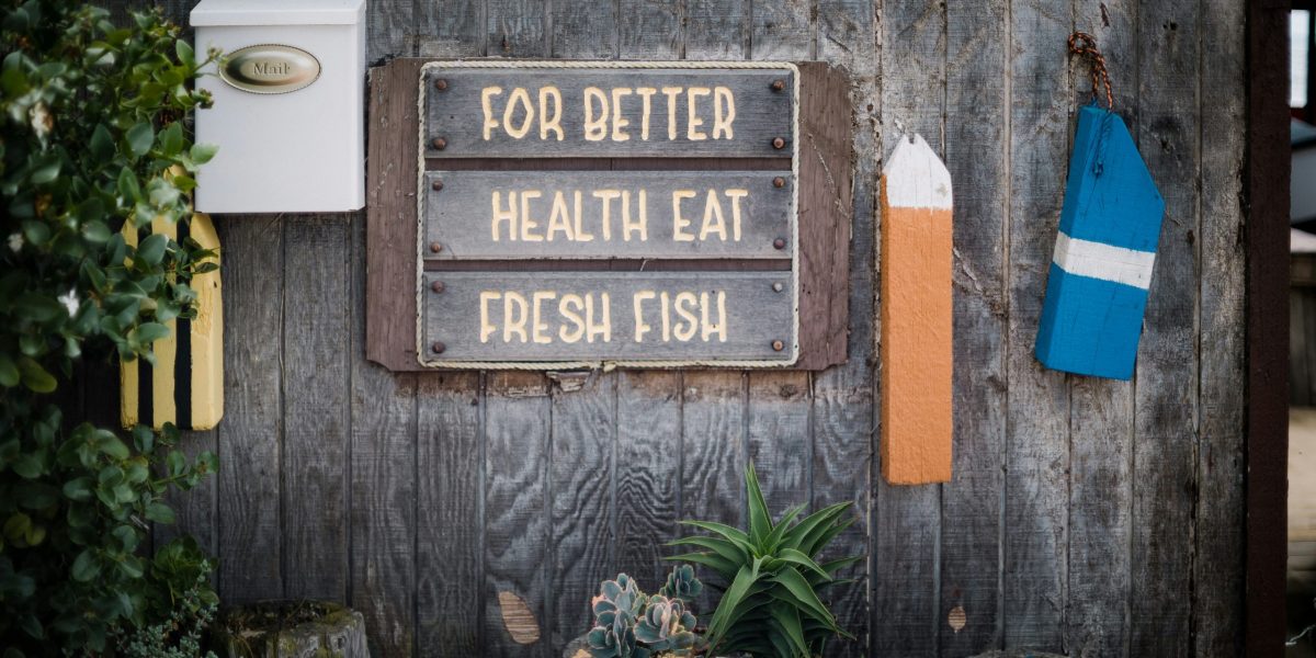 Mur de bois de grange sur lequel un panneau indique "Pour une meilleure santé, mangez du poisson frais". Une bouée bleue et orange, des plantes vertes et une boîte aux lettres blanche apparaît également sur ce mur.