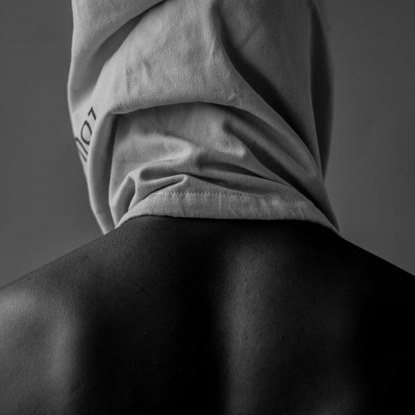 Personne de dos, torse nu, avec un sac en tissu sur la tête, dans une lumière noire et blanche, évoquant le mystère et l’introspection corporelle.