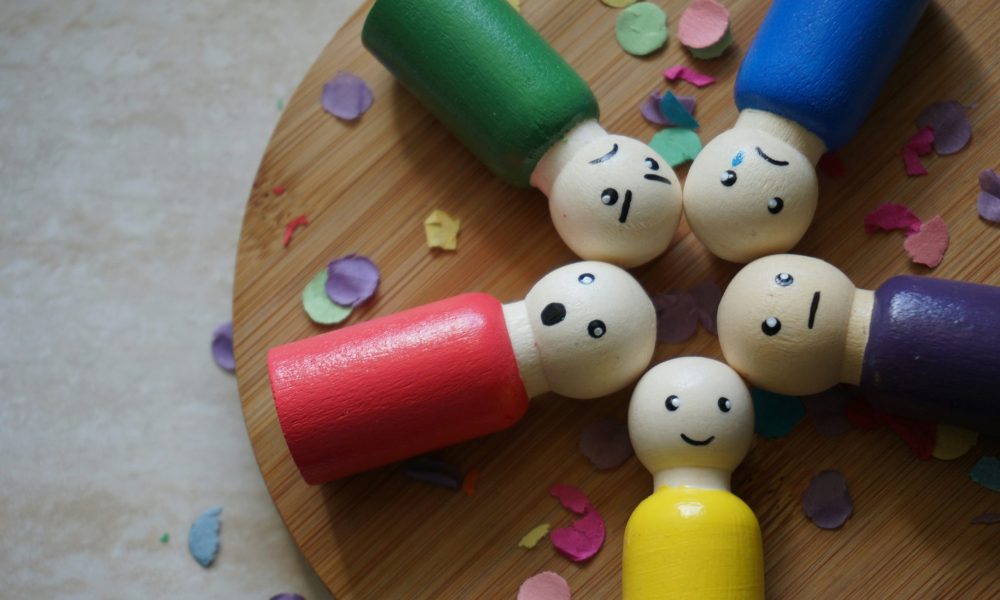 Cinq figurines en bois colorées formant un cercle, chacune exprimant une émotion différente, posées sur une surface en bois avec des confettis. Utilisée pour illustrer l'article abordant les impacts des émotions sur la santé.
