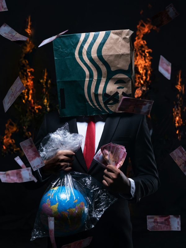 Homme en costume avec sac de marque sur la tête, tenant la planète emballée et des billets, avec des flammes en arrière-plan. Photo dénonçant les conséquences du mode de vie moderne.