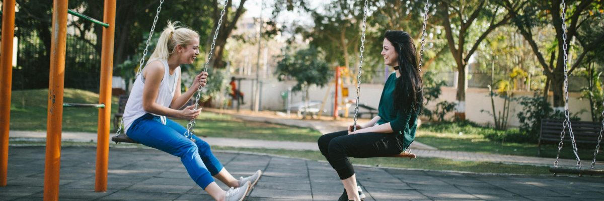 Deux amies souriantes en tenue de sport en pleine discussion sur des balançoires en léger mouvement dans un parc ensoleillé. Évocateur des bienfaits de bouger.